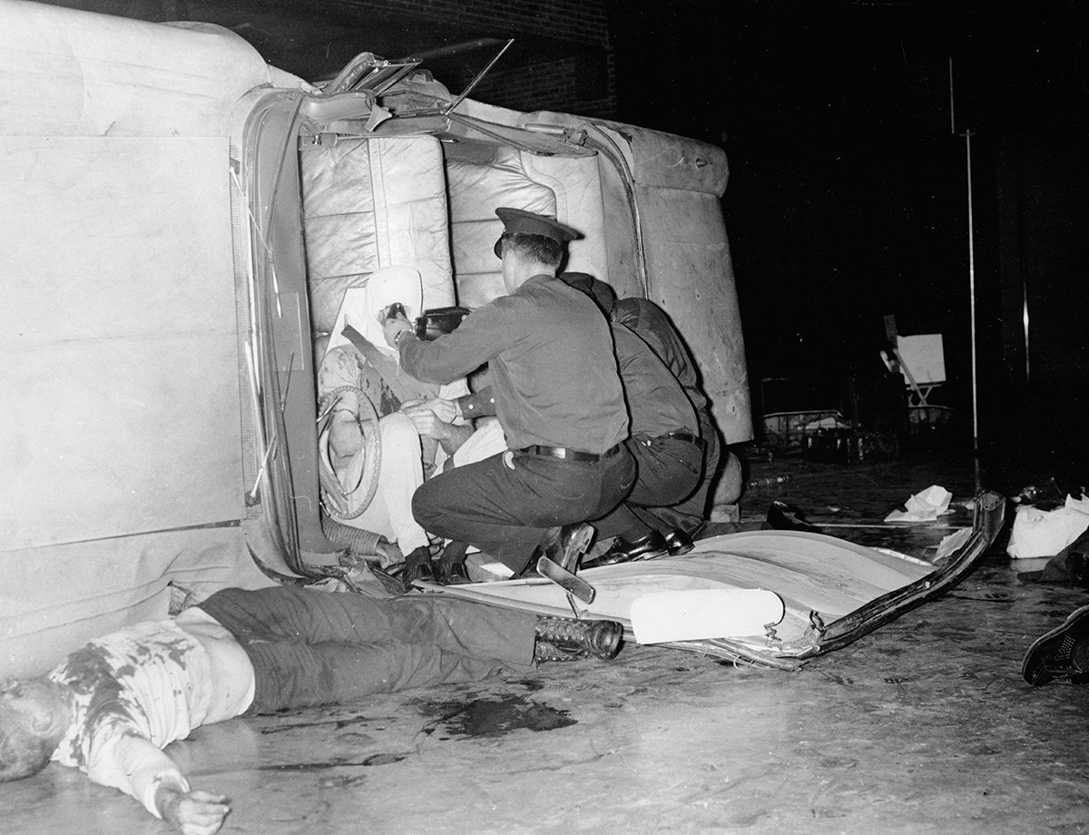 A photo showing a car on its side with emergency responders caring for the injured. 